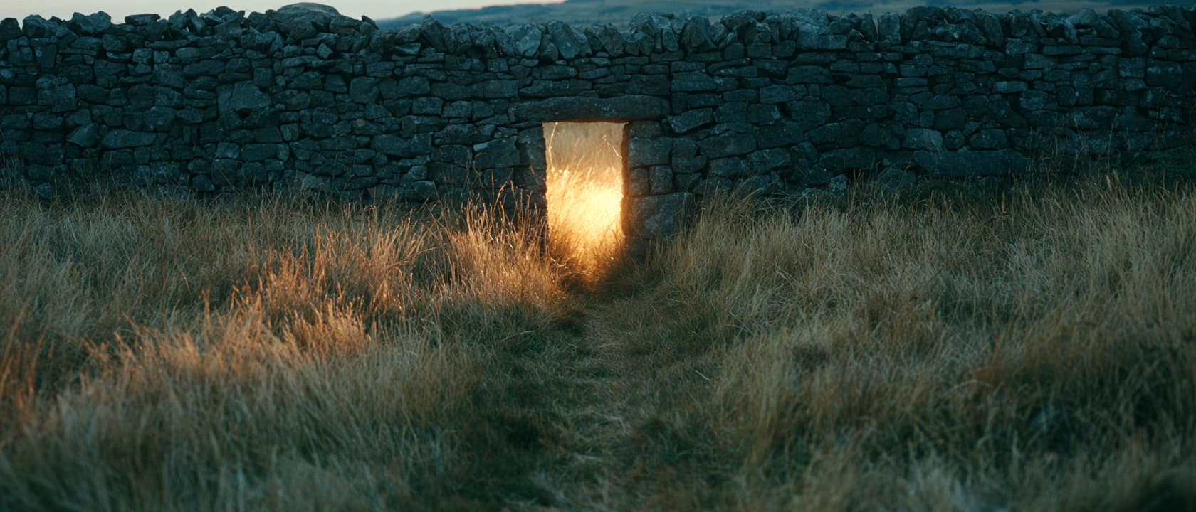 A worn path through tall grass leading to a doorway in an old stone wall, with warm light coming through the opening